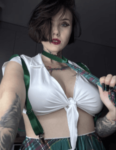 Self-portrait of a woman with short dark hair, wearing a white tied blouse, green suspenders, and a plaid skirt, holding a strap near her shoulder.