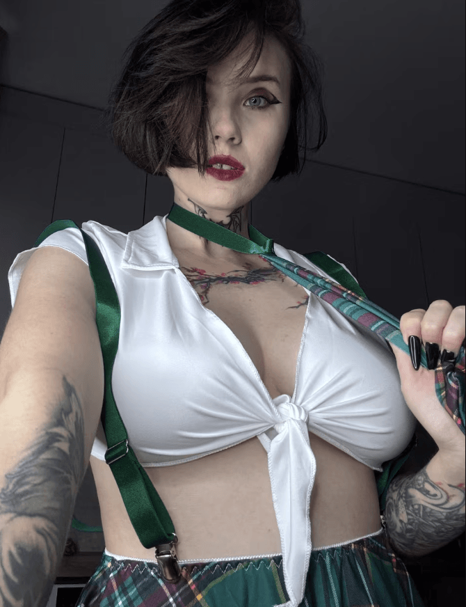 Self-portrait of a woman with short dark hair, wearing a white tied blouse, green suspenders, and a plaid skirt, holding a strap near her shoulder.