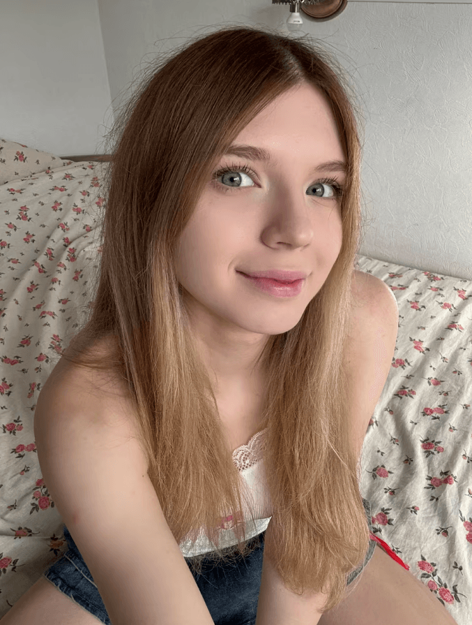 Young woman with long light brown hair and blue eyes smiling at the camera, seated on a floral bedspread.