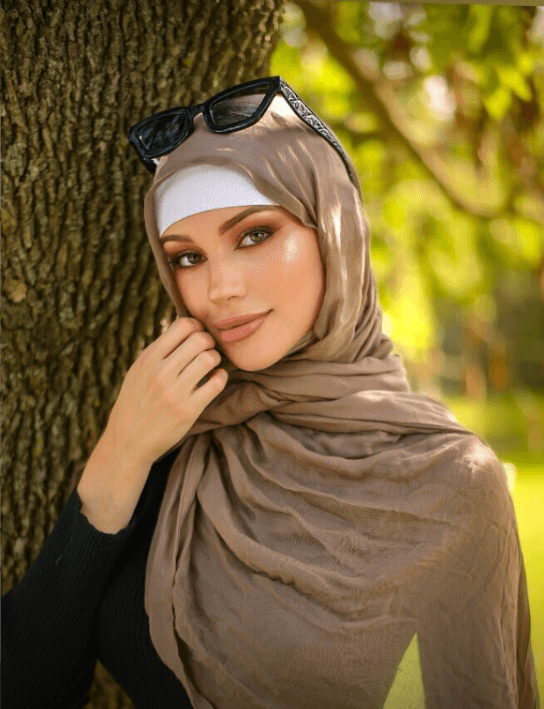 Woman wearing a beige hijab with a white undercap and sunglasses on her head, posing beside a tree in a sunlit park.