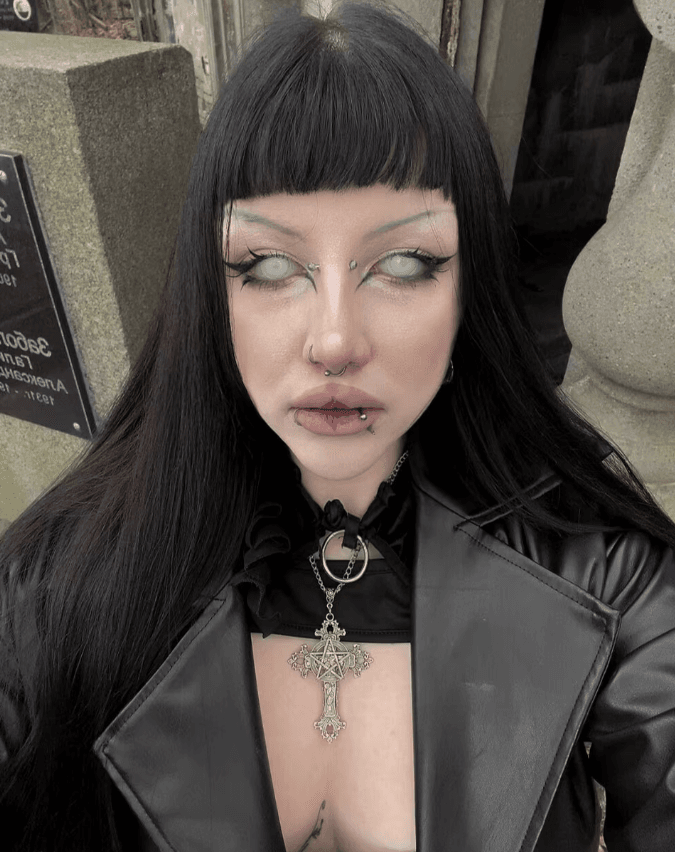 Person with black hair and bangs, wearing a black leather jacket and a large cross pendant.