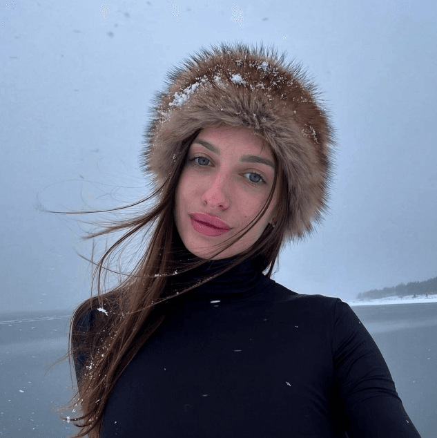 Young woman in a black turtleneck with a fur-lined hood in a snowy, windy landscape.