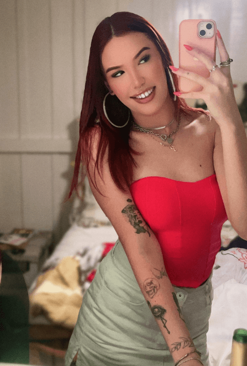 Woman taking a mirror selfie in a bedroom, wearing a red strapless top and green skirt, with tattoos on her left arm.