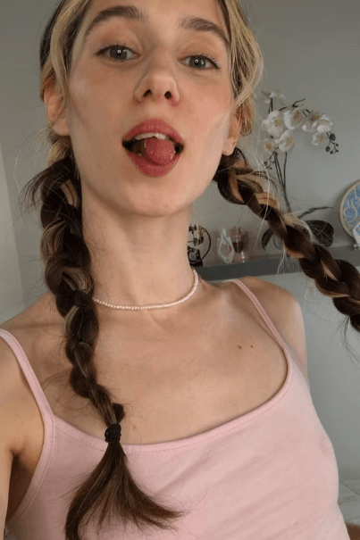 Close-up of a young woman with two braids, wearing a pink tank top and pearl necklace, sticking her tongue out playfully in a bright room.