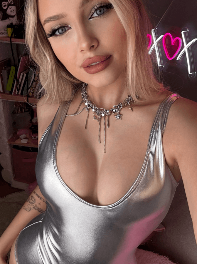 Close-up selfie of a woman with blonde hair wearing a shiny silver low-cut top and a starry choker necklace; pink neon signage in the background, tattoo visible on her left arm.