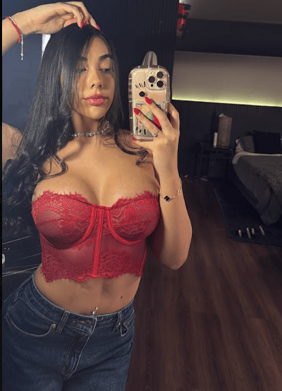 Woman taking a mirror selfie in a red lace bralette and jeans, in a modern bedroom backdrop.