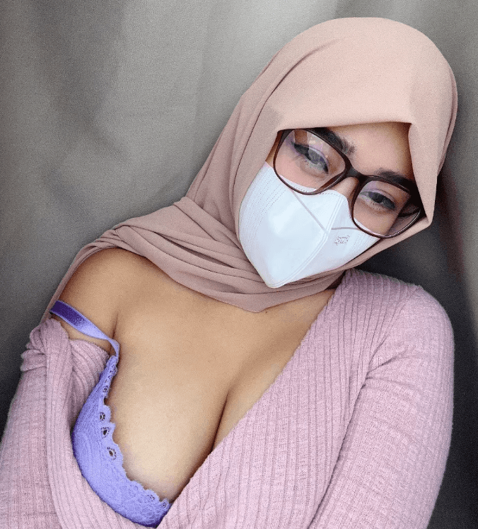 Close-up of a person wearing a beige hijab, white mask, and glasses, with a lavender lace bra visible under a pink cardigan