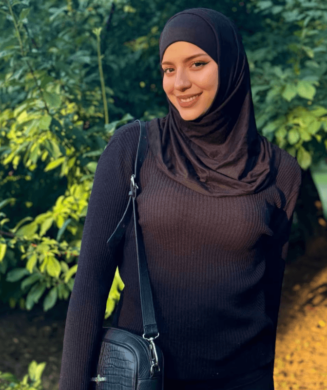 Woman in a black hijab and sweater stands outdoors among green bushes, smiling, with a black crossbody bag.