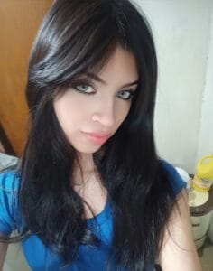 Close-up selfie of a young woman with long dark hair and a blue top, looking at the camera with a neutral background.