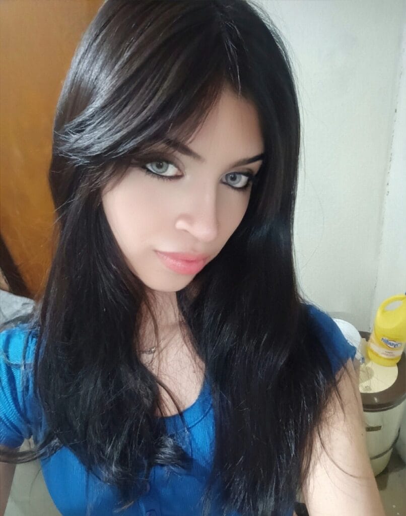 Close-up selfie of a young woman with long dark hair and a blue top, looking at the camera with a neutral background.