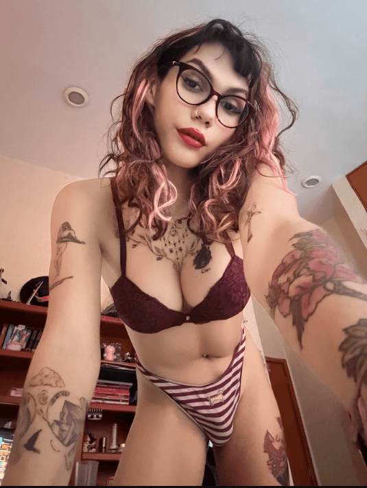 Selfie of a tattooed woman with pink-dyed hair and glasses wearing a burgundy lace bra and striped underwear in a bedroom.
