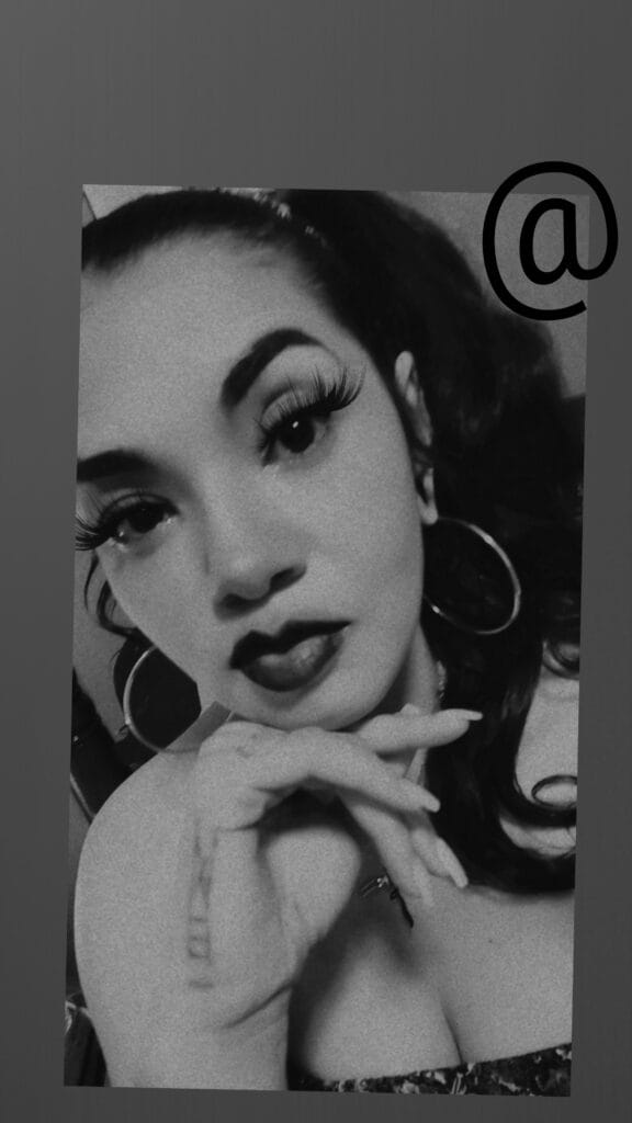 Black-and-white close-up portrait of a woman with bold makeup, hoop earrings, and hands resting under her chin.