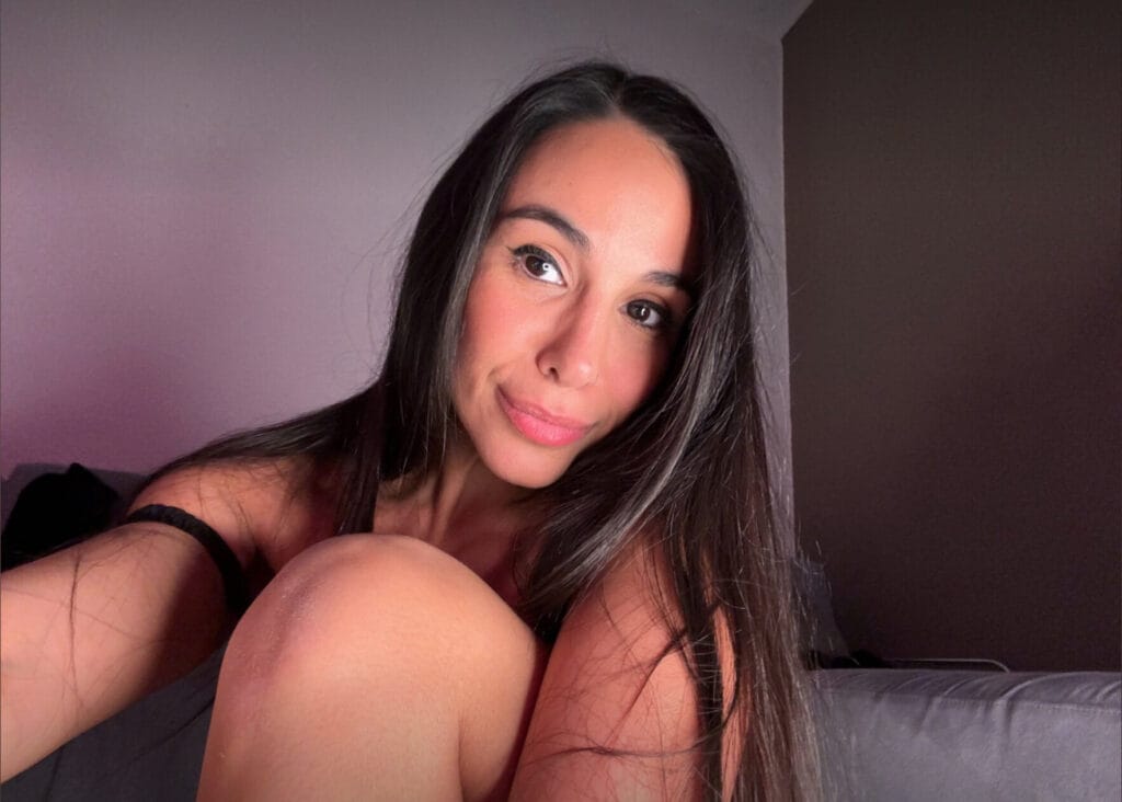 Woman taking a selfie, close-up of her face with long dark hair and pink lighting, sitting on a couch.