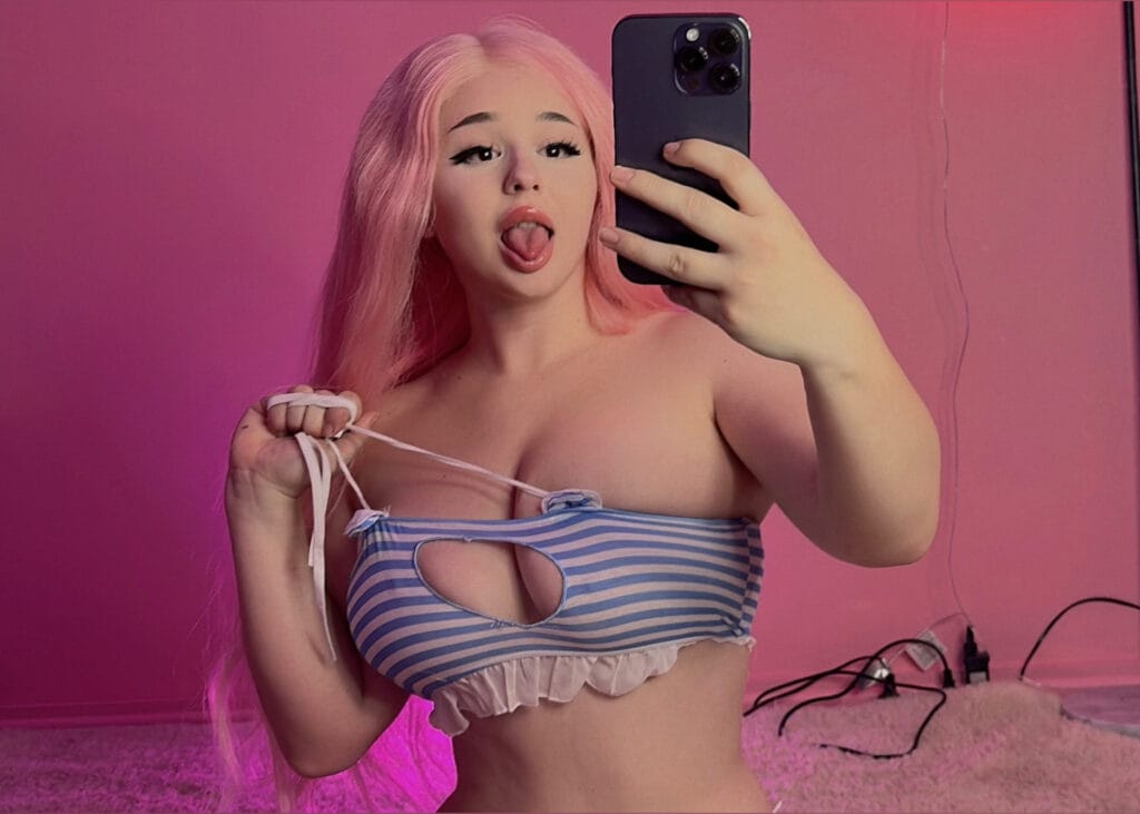 Person with pink hair taking a mirror selfie in a pink room, sticking out tongue and wearing a blue-and-white striped bikini top.