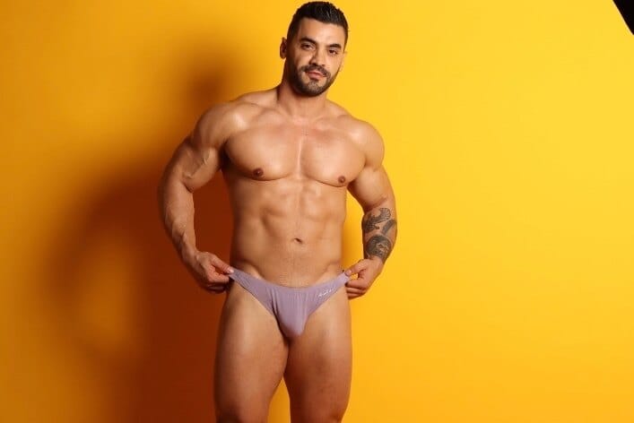 Muscular man in lavender briefs posing against a bright yellow backdrop in a studio setting.