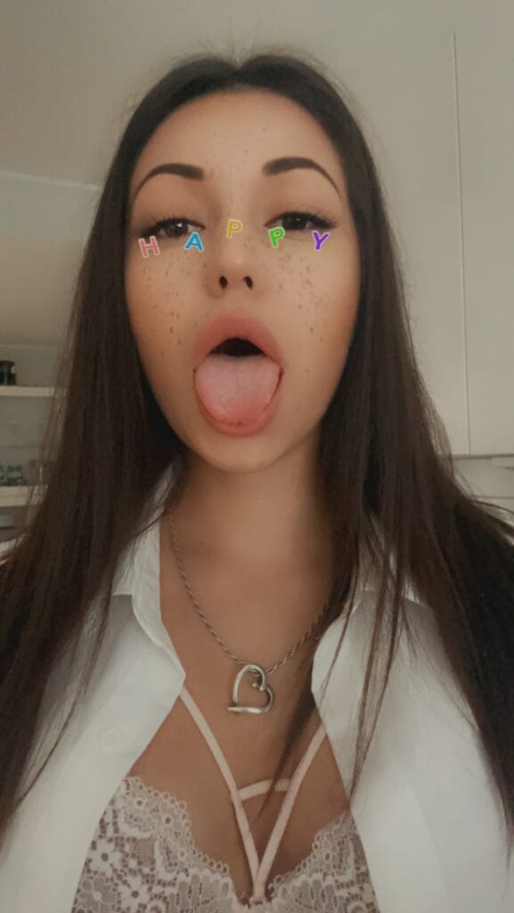 Young woman sticking out her tongue playfully with colorful HAPPY letters across her nose and freckles visible on her face, indoors in a white lace outfit and necklace.