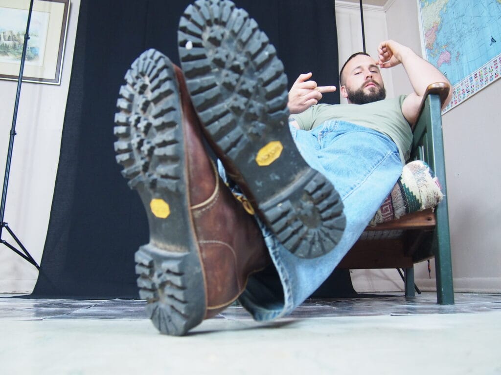 Low-angle shot of a bearded man lounging on a bench with boots raised toward the camera, wearing a green shirt and blue jeans; a world map is on the wall beside him.