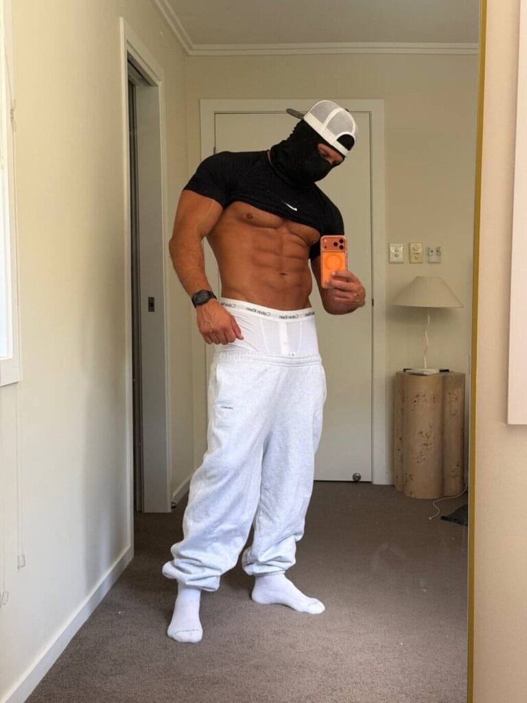 Mirror selfie of a person lifting a black shirt to reveal defined abs, wearing a white cap, white sweatpants, and a black mask in a bedroom bathroom area.
