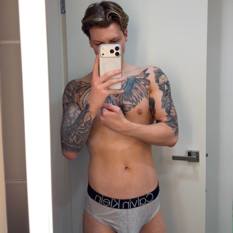 Shirtless man with large tattoos taking a mirror selfie, holding a beige iPhone and wearing gray Calvin Klein briefs in a bathroom door frame.