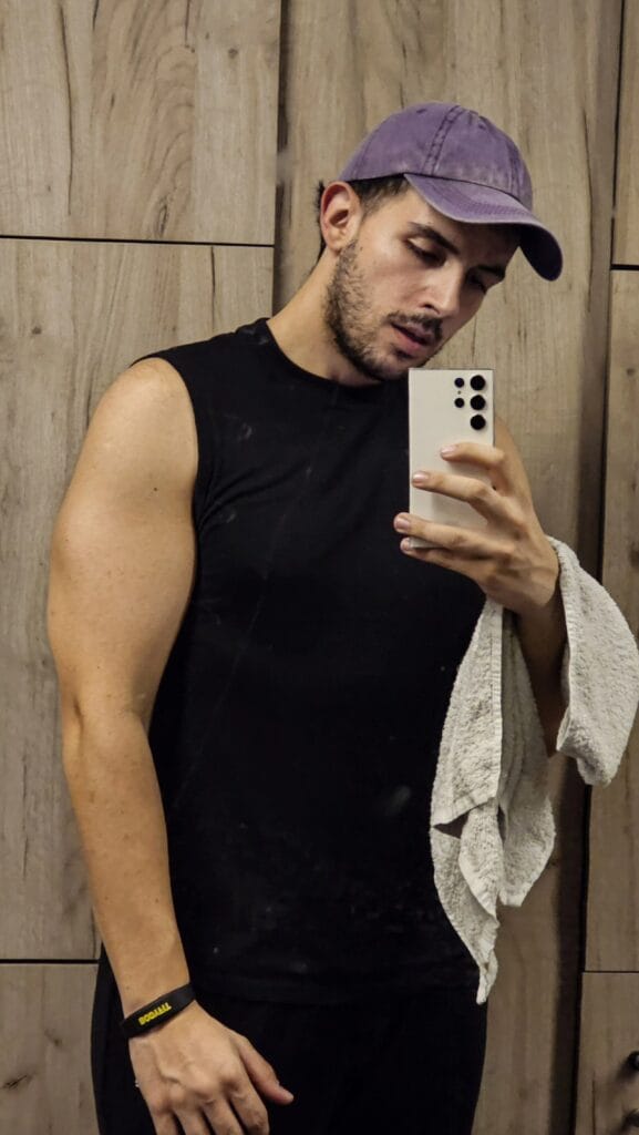 Young man wearing a purple cap and black sleeveless shirt takes a mirror selfie, holding a white phone with multiple cameras; towel draped over his arm in a wooden-panel room.