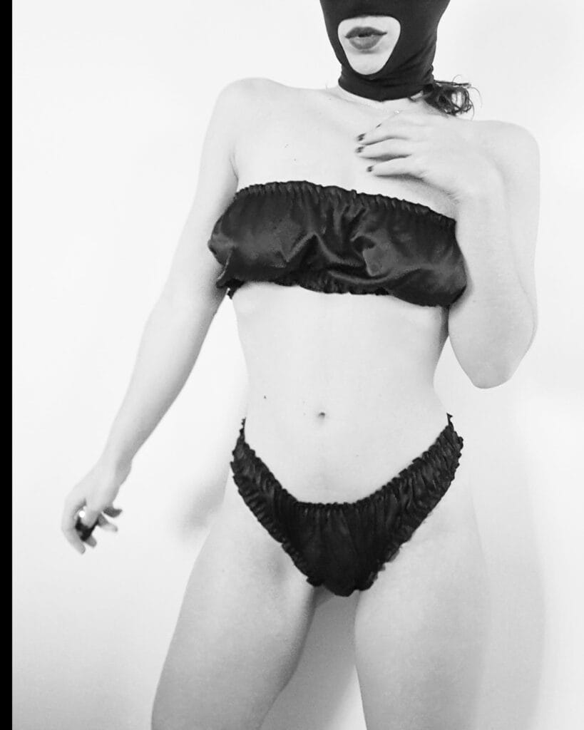 Black-and-white photo of a woman in a black bikini and a matching hood mask, standing in a studio.