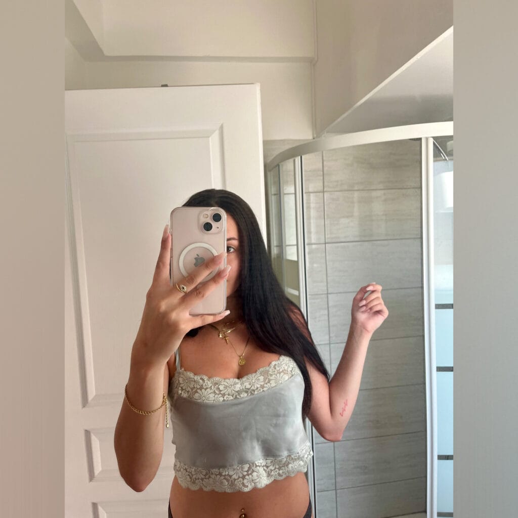 Woman taking a mirror selfie in a bathroom, wearing a light grey lace crop top and dark bottoms, holding a smartphone.